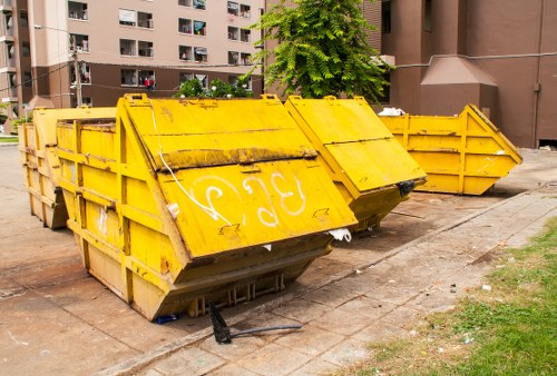 A yellow industrial skip with a metal frame and reinforced side panels, positioned on a concrete pavement in an urban residential area. The skip's exterior shows signs of weathering, with visible dirt and minor rust spots, and features white graffiti markings on the front. It is partially filled with various types of rubbish, with a black bag and some loose debris visible at the opening. The skip is placed next to a patch of grass and nearby there are two additional similar yellow skips, all aligned along the pavement. In the background, multi-storey residential buildings with balconies and windows are visible, along with a small tree and some greenery. The surrounding environment appears to be an apartment complex courtyard or communal outdoor space in Southwark, London. This scene is indicative of waste collection and rubbish disposal services provided by Garden Clearance Southwark, focusing on garden and waste clearance in a city setting.