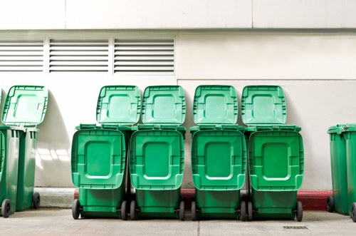 A row of eight green wheelie bins with closed lids is positioned along a paved outdoor surface adjacent to a modern building wall. The bins are uniform in size and design, with textured plastic surfaces and black wheels at the base, arranged in two equal rows of four. The background features a white wall with horizontal ventilation slats near the top, and a faint shadow cast by sunlight suggests a clear day. The bins are aligned against the wall, possibly awaiting collection, and are typical of waste management practices in Southwark and the surrounding London area. The overall scene reflects a maintained outdoor space ready for rubbish collection services, which Garden Clearance Southwark provides within the local postcode district.
