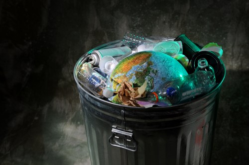 A close-up view of a metallic rubbish bin filled with various discarded items, including numerous empty plastic bottles, a crumpled brown paper bag, and other small plastic containers and packaging. The bin, which has a shiny silver finish and a secured lid latch, sits on a dark surface with a textured, possibly concrete or metallic background. The plastic bottles are clear, with some showing labels or nozzles, and are partially transparent, revealing their hollow interiors. The crumpled paper bag adds a contrasting brown tone amidst the shiny plastics, and the overall scene conveys the type of waste typically collected in a general rubbish removal service, consistent with the work of Garden Clearance Southwark in managing waste materials in Southwark and surrounding areas. The image highlights the common household or garden-related waste that requires professional clearance in the local area.
